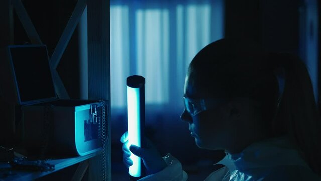 A female forensic expert removes fingerprints from a jewelry box with a brush. A woman collects evidence using an ultraviolet lamp at a crime scene in a dark apartment lit by red, blue police sirens.