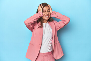 Teen girl in chic pink suit on a blue background laughs joyfully keeping hands on head. Happiness concept.