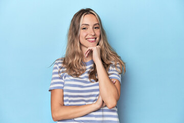 Blonde young caucasian woman in blue studio smiling happy and confident, touching chin with hand.