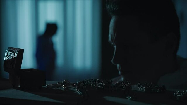 A male forensic investigator examines evidence, scattered jewelry, with a magnifying glass. A team of forensic examiners work at a crime scene in a dark apartment lit by blue red light.