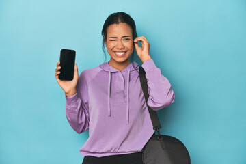 Filipina ready for gym with phone on blue covering ears with hands.