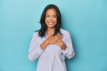 Filipina young woman on blue studio has friendly expression, pressing palm to chest. Love concept.