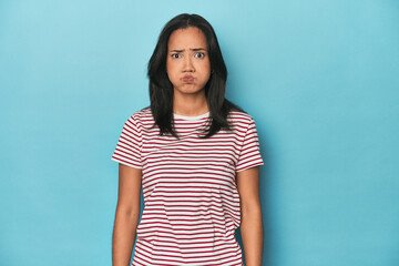 Filipina young woman on blue studio blows cheeks, has tired expression. Facial expression concept.