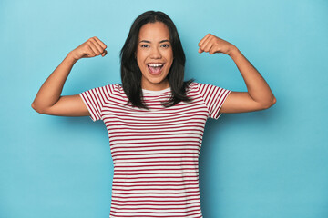 Filipina young woman on blue studio showing strength gesture with arms, symbol of feminine power