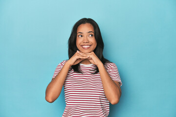 Fototapeta premium Filipina young woman on blue studio keeps hands under chin, is looking happily aside.
