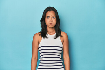 Filipina young woman on blue studio blows cheeks, has tired expression. Facial expression concept.