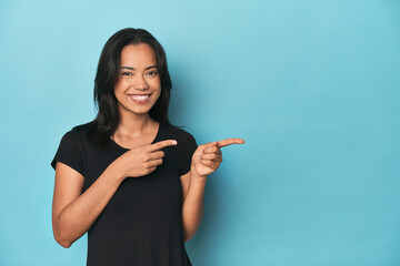 Filipina young woman on blue studio excited pointing with forefingers away.