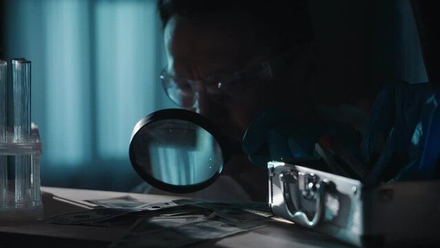 A forensic investigator using a magnifying glass examines dollar bills with drops of blood. A man takes a blood sample from a banknote and applies it to a slide.