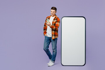 Full body young man wears checkered shirt white t-shirt casual clothes stand near big huge blank screen mobile cell phone with area using smartphone isolated on plain pastel light purple background.