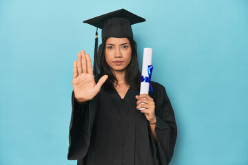 Filipina graduate with diploma on blue studio standing with outstretched hand showing stop sign,...