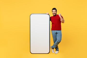 Full body young Indian man wear red t-shirt casual clothes big huge blank screen mobile cell phone smartphone with area do winner gesture isolated on plain yellow orange background. Lifestyle concept