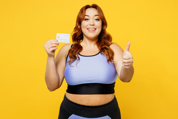 Young fun chubby overweight plus size big fat fit woman wear blue top warm up training hold credit bank card show thumb up isolated on plain yellow background studio home gym. Workout sport concept.
