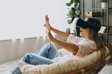 Side view young fun woman wear casual clothes sits in armchair watching in vr headset pc gadget stay at home hotel flat rest relax spend free spare time in living room indoor Lifestyle lounge concept.