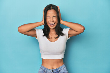 Filipina young woman on blue studio covering ears with hands trying not to hear too loud sound.