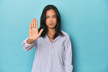 Filipina young woman on blue studio standing with outstretched hand showing stop sign, preventing...