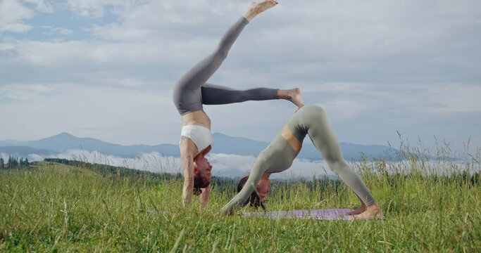Active caucasian women in sport clothes doing acro yoga together on fresh air. Two female athletes balancing on yoga mat. Wellness and wellbeing concept