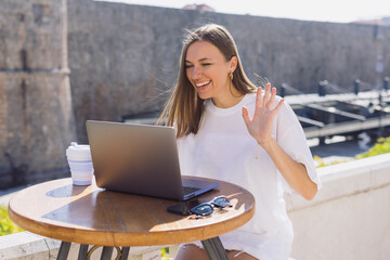 A businesswoman waves her hand into a laptop camera during a call with business partners. A woman is sitting outdoors in a cafe - obrazy, fototapety, plakaty