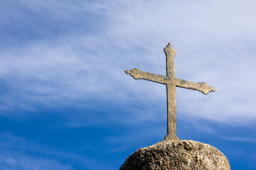 Cruz católica metálica sobre pedestal de roca, con un cielo azul con nubes de fondo. Espacio de...