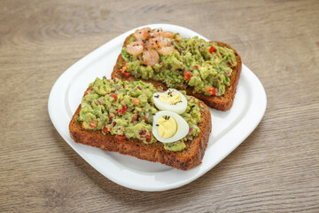 Slices of bread with tasty guacamole, eggs and shrimp on wooden table, closeup