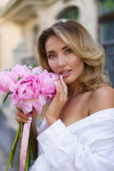 Elegant attractive blond woman holding amazing pink bouquet with pions.  Walking  outdoor in european city.  Wearing casual summer outfit, white blouse, silk skirt.