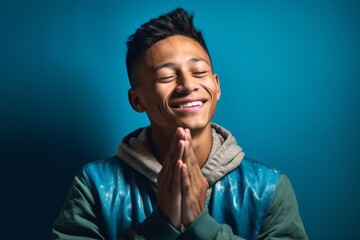 Close-up portrait photography of a grinning boy in his 20s putting hands together as if praying against a teal blue background. With generative AI technology