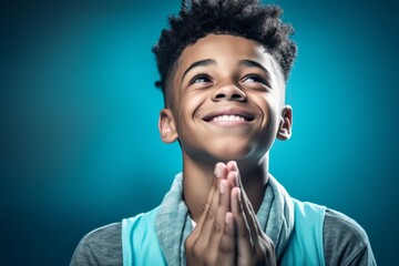 Close-up portrait photography of a grinning boy in his 20s putting hands together as if praying against a teal blue background. With generative AI technology
