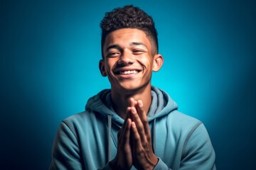 Fototapeta premium Close-up portrait photography of a grinning boy in his 20s putting hands together as if praying against a teal blue background. With generative AI technology