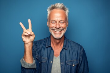 Lifestyle portrait photography of a grinning mature man making a peace and love gesture with the fingers against a soft blue background. With generative AI technology
