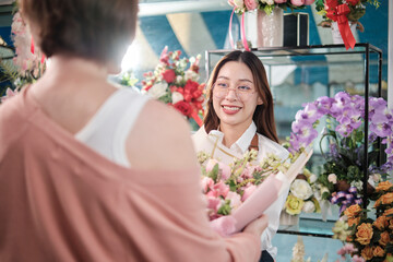 Young Asian female florist worker in apron delivers beautiful fresh blossoms bouquet to customer who purchased order, happy seller who works in colorful flower shop, and small business entrepreneur.