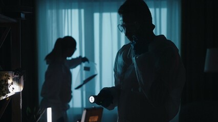 Forensic specialists work at the crime scene, in a dark apartment lit by blue light from police sirens. A man with a flashlight speaks on his smartphone, while a woman examines the murder weapon.