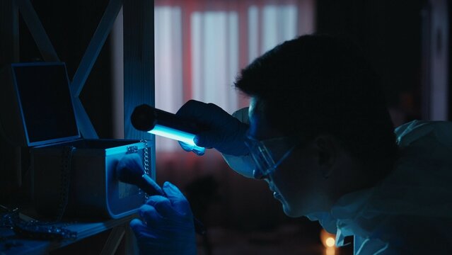 A male expert criminologist removes fingerprints from a jewelry box with a brush. A man gathers evidence using an ultraviolet lamp at a crime scene in a dark apartment lit by red, blue police sirens.