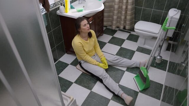High angle view wide shot of exhausted young woman sitting on floor in bathroom at home. Portrait of tired Caucasian attractive housewife taking break cleaning house indoors