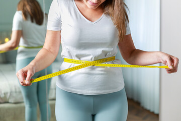 Young woman measuring her waist with tape measure in front of the mirror in her bedroom
