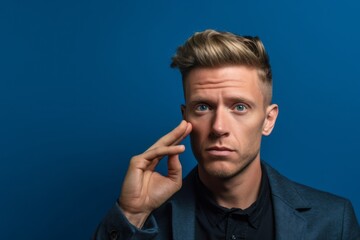 Close-up portrait photography of a beautiful boy in his 30s making a gesture of i'm thinking with the finger on the head against a sapphire blue background. With generative AI technology