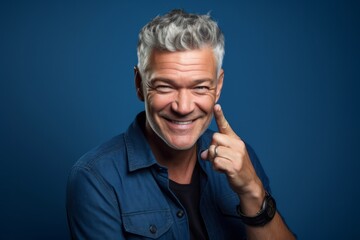 Lifestyle portrait photography of a grinning mature man making a gesture of i'm thinking with the finger on the head against a sapphire blue background. With generative AI technology