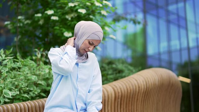 Tired young muslim female employee in hijab suffering from neck pain while sitting on a bench on the street near an office building. Sad woman massages and rubs sore muscles with her hand, stretches
