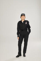 Obraz na płótnie Canvas full length of smiling policewoman with pink ribbon on uniform on grey background, breast cancer