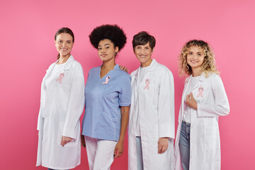 Obraz premium smiling interracial doctors with ribbons looking at camera isolated on pink, breast cancer concept