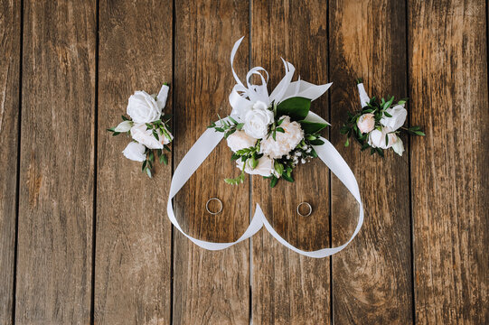 A bouquet of flowers with a ribbon in the form of a heart, a boutonniere, golden rings of the newlyweds lie on a wooden background. Close-up wedding photo, top view.