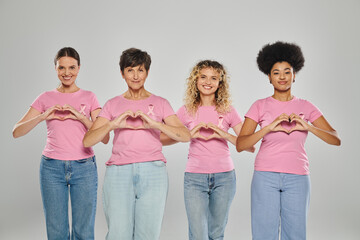 Fototapeta premium breast cancer awareness, happy interracial women with pink ribbons showing heart on grey, diversity