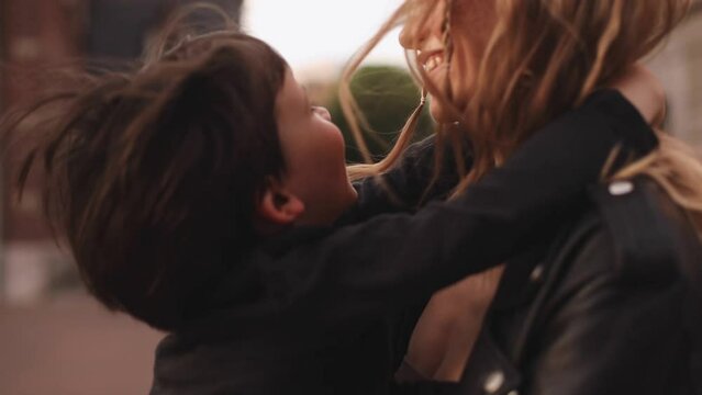 Attractive blonde curly woman with two thin braids wear black leather jacket hold her brunette son in long sleeves t-shirt and turn around him, spinning, boy show his tongue, they happy. 