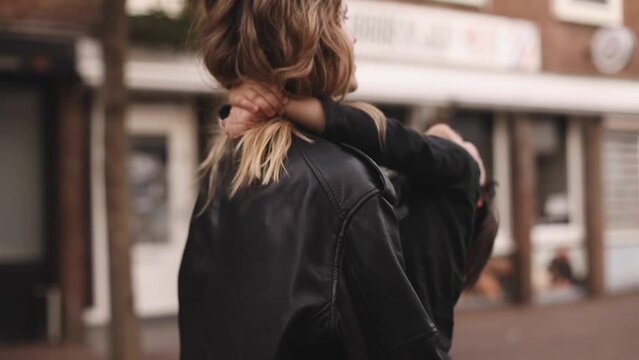 Attractive blonde curly woman with two thin braids wear black leather jacket and dress carries in her arms her brunette son, he hugs mothers neck, and touch her hairs, family go home. Selective focus.