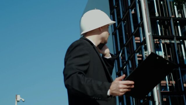 Male civil engineer contractor in safety helmet and jacket standing on construction site building and using mobile phone talking discussion real estate development working plan with architect manager.