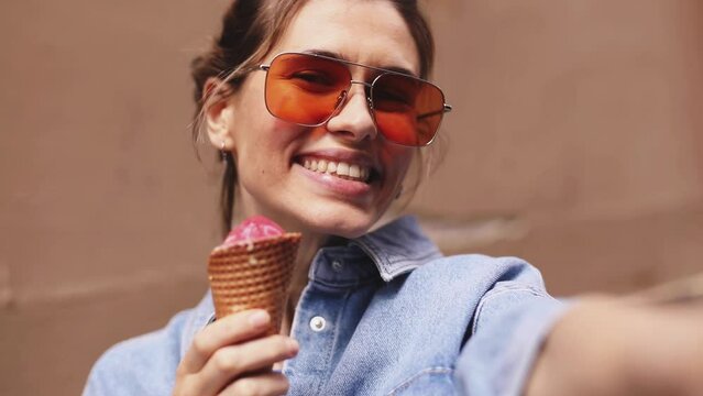 Laughing beautiful young blonde woman with high ponytail posing, walking on summer street outdoors take selfie by mobile phone eat ice cream. Attractive woman wear orange sunglasses, make photo.