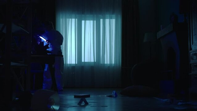 A team of forensic specialists work at the crime scene, in a dark apartment lit by blue red light from police sirens. A man and a woman using ultraviolet lamps are looking for evidence, fingerprints.