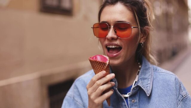 Close up portrait woman with high ponytail hold delicious pink ice cream in waffle cone outdoors. Girl wear orange sunglasses. Female hold ice cream, bite, lick and walking by the street, enjoy