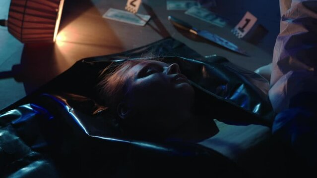 A murdered woman in a dark apartment illuminated by blue red light from police sirens close up. The forensic expert hands are packing the victim's body into a body bag.