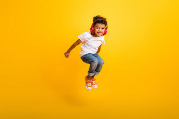 Fototapeta na wymiar Full length photo of carefree excited small boy wear white t-shirt jumping high listening music headphones isolated yellow color background