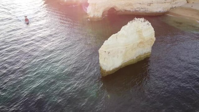 Aerial Drone Reverse Shot of Ocean, Rock, Boat, and Beach. A boat cruises beside a rock in the ocean, while the beach lies to the right. Sun rays shine throughout. 