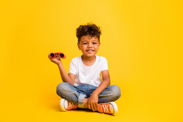 Full length photo of sweet cheerful small boy wear white t-shirt rising binoculars isolated yellow...